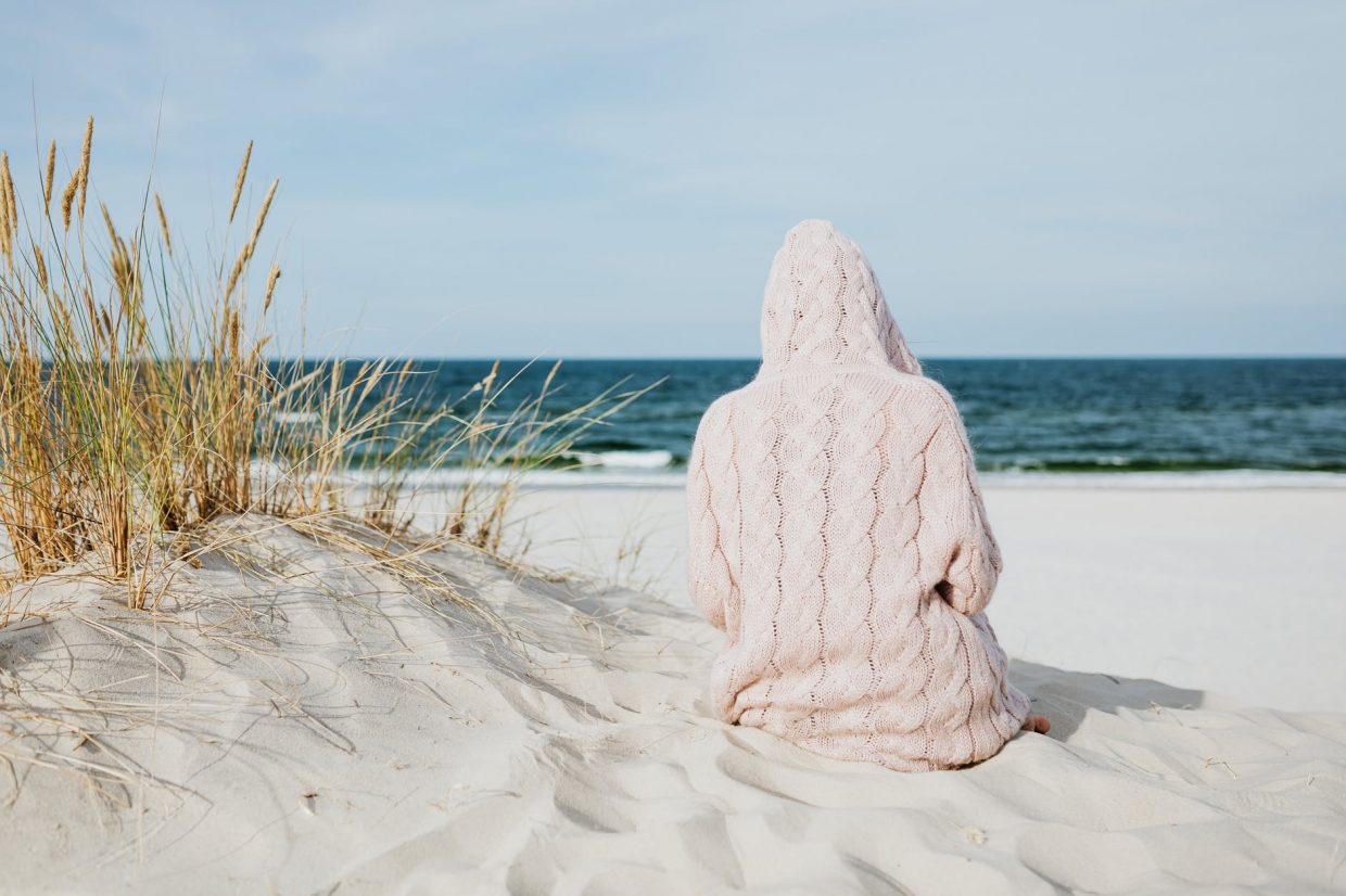 person wearing hoodie sitting on white sand near body of water